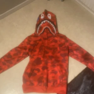 Röd camo hoodie med hajmotiv - Cool röd hoodie med camouflagemönster och hajmotiv på huvan. Har dragkedja och unika detaljer som bokstäver och grafik på sidan av huvan. Perfekt för en streetwear-look.