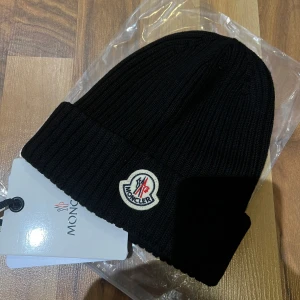 Svart ribbad mössa från Moncler - Snygg svart ribbad mössa från Moncler med broderad logotyp framtill. Perfekt för att hålla värmen under kyliga dagar. Klassisk design som passar till det mesta.