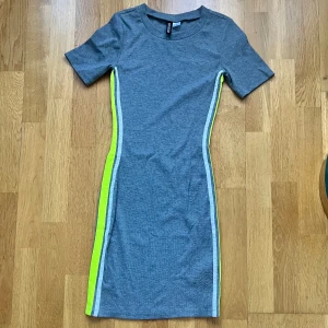 Grå klänning med neongula detaljer - Snygg grå klänning med korta ärmar och neongula ränder längs sidorna. Perfekt för en sportig look med en figurnära passform. Klänningen är tillverkad i ett mjukt och stretchigt material för extra komfort.