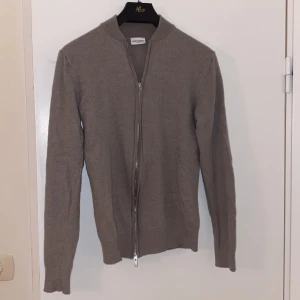 Beige merinoullkofta från John Henric - Säljer en stilren beige kofta från John Henric i 100% merinoull. Koftan har en dragkedja framtill och långa ärmar, perfekt för kyligare dagar. Passar både till vardags och mer uppklädda tillfällen. Modellen är 181 och väger 68 kg✅