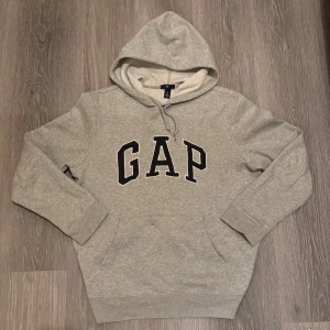Grå hoodie från GAP - Säljer en klassisk grå hoodie från GAP med stor logga på bröstet. Tröjan har en bekväm huva och en praktisk magficka. Perfekt för en avslappnad stil. Skriv för mer info:)
