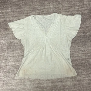 Vit blus med spetsdetaljer - Säljer en söt vit blus med v-ringning och spetsdetaljer. Blusen har korta ärmar och ett fint hålmönster som ger en romantisk känsla. Perfekt för varmare dagar eller som ett lager under en kavaj.