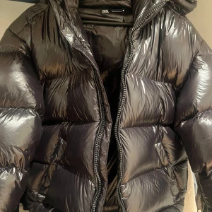 Svart pufferjacka från Zara - Snygg svart pufferjacka från Zara med glansig finish och dragkedja framtill. Jackan har en huva.  Är köpt på Zara herravdelning till sonen men är unisex. Endast använd 1 gång och säljes pga fel stl.  Ord pris 1395kr 