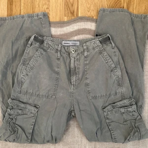 Grå cargobyxor från Bershka - Säljer ett par grå cargobyxor från Bershka. Byxorna har flera praktiska fickor och en loose passform. Perfekta för en avslappnad stil. De har en knapp och dragkedja framtill samt bälteshällor.