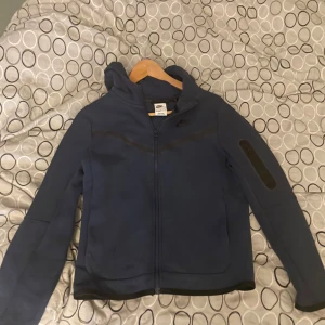 Nike tech fleece  - Säljer en mörkblå jacka från Nike med dragkedja och huva. Jackan har en stilren design med en ficka på ärmen och är perfekt för vardagsbruk. 