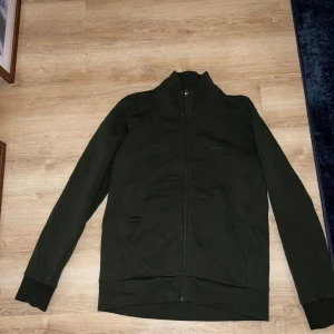 Grön tröja med dragkedja från Boss - Snygg grön tröja från Boss med dragkedja och hög krage. Perfekt för en stilren look. Tröjan har långa ärmar och ribbade muddar vid ärmslut och nederkant.