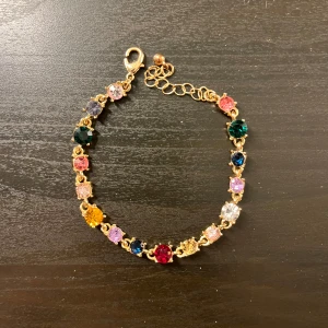 Färgglatt armband med stenar - Snyggt armband med en rad av färgglada stenar i olika nyanser som röd, blå, grön och rosa som liknar Caroline Svedbom. Armbandet har en justerbar kedja och en guldfärgad bas. Perfekt för att lägga till en färgklick till din outfit!
