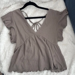 Beige blus med volangdetaljer - Säljer en söt beige blus med volangdetaljer och v-ringning. Blusen har korta ärmar och knytning i ryggen. Den är sååå fin men har tyvärr inte kommit till användning så den är i nytt skick!🌸🌸💕💕