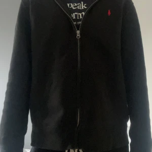 Ralph Lauren hoodie - Säljer en svart hoodie från Ralph Lauren med dragkedja och röd broderad logga på bröstet. Den har långa ärmar och en bekväm passform. Perfekt för en avslappnad stil.