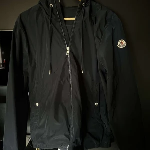 Moncler jacka - Snygg svart vindjacka från Moncler med dragkedja och huva. Jackan har långa ärmar och ett stilrent märke på ärmen. Perfekt för blåsiga dagar med sin lätta och funktionella design.
