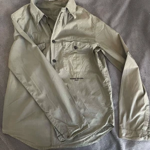Stone Island skjorta - Snygg beige skjorta från Stone Island med klassisk krage och knappar framtill. Skjortan har två bröstfickor med knappar och broderad logotyp på ryggen. Färgen matchar med det mesta och kan aldrig gå fel med en riktigt fin skjorta! Vid minsta lilla fundering kontakta oss!/ R.EsellingSWE😎