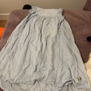 Ljusblå klänning från Pompdelux - Charmig ljusblå klänning från Pompdelux med volangdetaljer vid ärmarna. Perfekt för varma dagar med sin luftiga design. Klänningen har en avslappnad passform och är tillverkad i ett mjukt material.