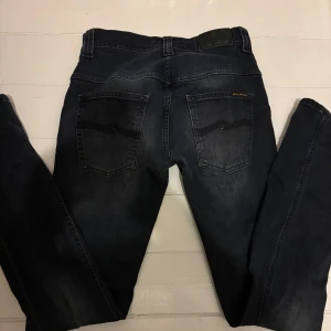 Mörkblå jeans från Nudie Jeans - Snygga mörkblå jeans från Nudie Jeans med klassisk femficksdesign och knappgylf. Perfekta för en stilren look. Märkesdetaljer syns på bakfickan och midjebandet.