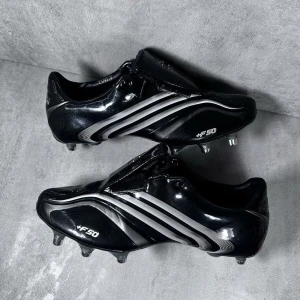 Svarta fotbollsskor Adidas F50 - Snygga svarta fotbollsskor från Adidas F50-serien med vita detaljer och klassiska ränder. Skorna har dobbar för bra grepp på planen och en stilren design.