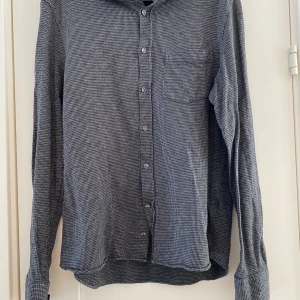 Grå rutig skjorta från J.Lindeberg - Snygg grå rutig skjorta från J.Lindeberg med klassisk krage och knappar framtill. Skjortan har en diskret bröstficka och är perfekt för en stilren look. Tillverkad i ett mjukt material som känns skönt mot huden. Ny grå och svart rutig skjorta i strl M från J.Lindeberg, modell Jersey Houndstooth slim shirt.  Ny, aldrig använd.  Mått Över bröstet: ca 51 cm Längd från axeln och ner: ca 67 cm  100% bomull  Kommer från ett djur och rökfritt hem. 