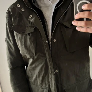 J.Lindeberg fieldjacket - Säljer en grön fieldjacket med dragkedja och knappar. Jackan har flera fickor framtill och en hög krage. Perfekt för höst och vår med sin stilrena design och praktiska detaljer.