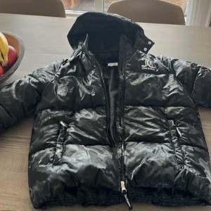 Svart pufferjacka från Juicy Couture - Säljer en snygg svart pufferjacka från Juicy Couture med ett diskret mönster och logga på bröstet. Jackan har en dragkedja framtill och en huva för extra skydd mot väder. Perfekt för kyliga dagar!