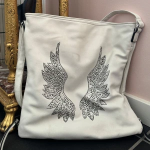 Vit axelväska med vingdetaljer - Snygg vit axelväska med glittriga vingdetaljer på framsidan. Väskan har dragkedja på  sidorna och en på baksidan för extra förvaring. 