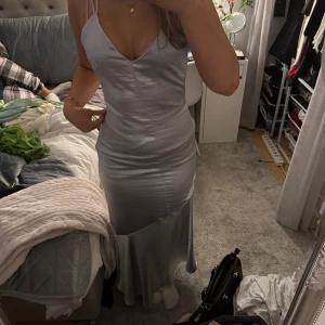 Silverfärgad långklänning med smala axelband - Elegant silverfärgad långklänning med smala axelband och v-ringning. Klänningen har en figurnära passform och en glansig yta som ger en lyxig känsla. Perfekt för speciella tillfällen. Har använts en gång på 9ans bal.