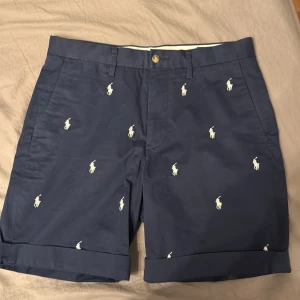 Mörkblå shorts från Polo Ralph Lauren - Snygga mörkblå shorts från Polo Ralph Lauren med broderade vita logotyper över hela tyget. Shortsen har en klassisk passform med knapp och dragkedja framtill. Perfekta för en stilren look. Dom är nästan aldrig använda köpta i usa nypris ca 2000