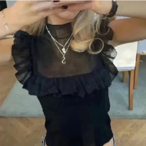 Svart transparent topp med volangdetaljer - Snygg svart transparent topp med volangdetaljer runt axlarna och bröstet. Perfekt för en trendig look. Toppen har en tight passform och är ärmlös, vilket gör den idealisk för varmare väder eller lager-på-lager-stil. Lånade bilder!!!