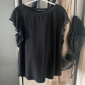 Svart blus från Zara  - Säljer en elegant svart blus från Zara Woman. Blusen har korta ärmar med volangdetaljer som ger en fin touch. Perfekt för en stilren look. Jättefin och passar till allt. Inga defekter
