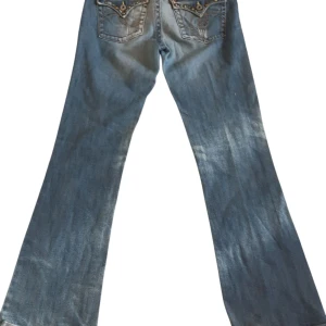 Blå jeans med nitar och broderi - Snygga blå jeans med bootcut-stil och coola detaljer som nitar och broderi på bakfickorna. De har en klassisk femficksdesign och en knappgylf. Perfekta för en avslappnad look med en twist.