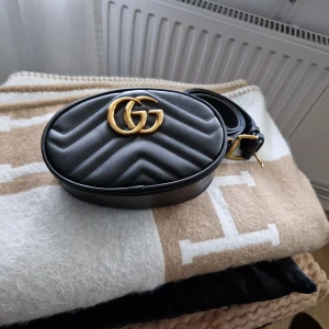 Svart quiltad midjeväska med gulddetaljer - Snygg svart quiltad midjeväska med ett hjärtmönster på baksidan och guldfärgad logga framtill. Väskan har en justerbar rem och dragkedja för säker förvaring. Perfekt för att bära det viktigaste med stil.