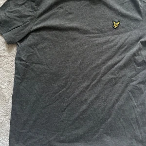 Grå t-shirt från Lyle & Scott - Säljer en stilren grå t-shirt från Lyle & Scott med deras ikoniska gula logga på bröstet. Perfekt för en avslappnad look. T-shirten är kortärmad och gjord i mjuk bomull.