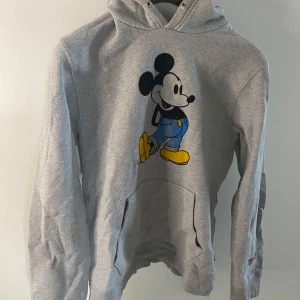 Grå hoodie med Musse Pigg från Levis - Grå hoodie från Levi's med en stor Musse Pigg-tryck på framsidan. Tröjan har en klassisk huva och magficka och touch av nostalgi. 🐭