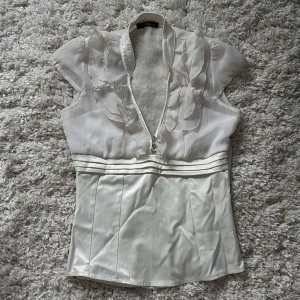 Vit topp med volanger - Superfin benvit topp från Jane Norman med v-ringning samt volangdetaljer upptill och fina knappar framtill. ✨Den har en dragkedja vid sidan. Otroligt vacker topp men aningen för tight för mig! Skriv om du har funderingar eller vill ha mått 💞