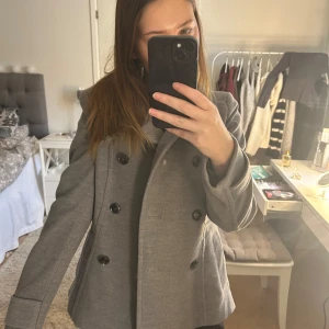 Grå kappa med knappar - Snygg grå kappa med dubbelknäppning och hög krage. Klassisk design som funkar till det mesta. Helt nyskick så ej använd