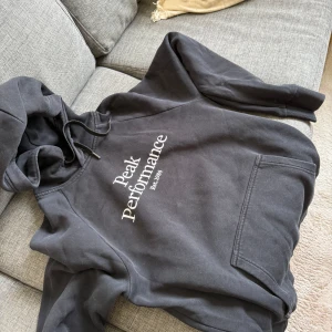 Svart hoodie från Peak Performance - Säljer en svart hoodie från Peak Performance med tryck framtill. Den har en klassisk design med huva och en stor ficka på magen. Perfekt för en avslappnad stil.
