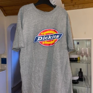 Grå t-shirt från Dickies - Säljer en grå t-shirt från Dickies med deras ikoniska logga i rött, gult och blått på bröstet. T-shirten är kortärmad och tillverkad i bomull, perfekt för en avslappnad stil. Passar bra till jeans eller shorts för en casual look. Skavanker på loggan 