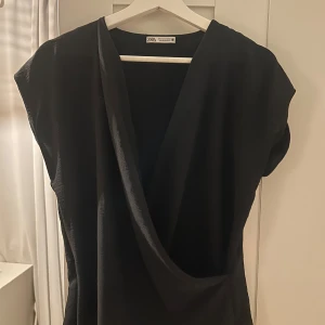 Svart omlott topp från Zara - Snygg svart omlott topp från Zara med en djup v-ringning. Perfekt för en stilren look. Den har korta ärmar och en elegant drapering framtill. Sitter mer som en S än M