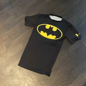 Svart Batman t-shirt från Under Armour - Säljer en svart kompressionst-shirt från Under Armour med det ikoniska Batman-logotypen i gult på bröstet. Perfekt för träning eller som en cool vardagst-shirt. Kortärmad design med HeatGear-teknologi för att hålla dig sval och bekväm.