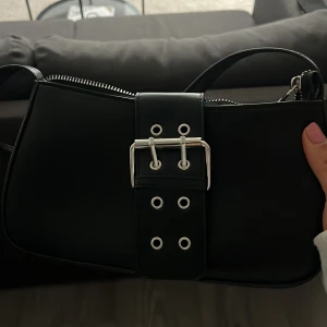 Helt oanvänd Svart axelväska med bälte - Snygg svart axelväska med coolt bältesdetalj framtill. Väskan har en dragkedja upptill och en smidig axelrem. Perfekt för att ge din outfit en edgy touch