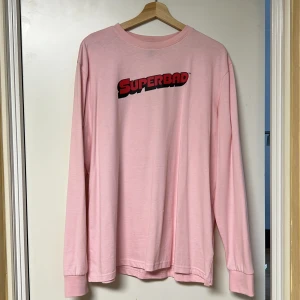 Rosa långärmad tröja med Superbad-tryck - Säljer en rosa långärmad tröja med Superbad-tryck på både fram- och baksidan. Tröjan har en avslappnad passform och är perfekt för fans av filmen. Den är gjord i ett mjukt material som känns skönt mot huden.