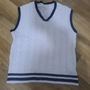 Vit stickad väst med svarta detaljer - Säljer en stilren vit stickad väst med svarta detaljer vid ärmhål och nederkant. Västen har ett klassiskt flätmönster och en v-ringad hals. Perfekt för en preppy look!