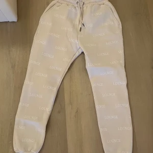 Beige mjukisbyxor från lounge - Säljer ett par beige mjukisbyxor med ordet 'LOUNGE' tryckt över hela byxorna. De har en elastisk midja med snörning för justerbar passform. Perfekta för en avslappnad stil.