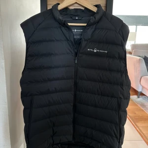 Sail Racing väst - Snygg svart dunväst från Sail Racing med dragkedja framtill. Perfekt för kyligare dagar och ger en vardaglig look. Västen har en stilren design med logotyp på bröstet.