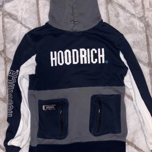Blå och grå hoodie från Hoodrich - Säljer en snygg blå och grå hoodie från Hoodrich med vit text på bröstet. Den har två praktiska fickor med dragkedjor framtill och en justerbar huva. Perfekt för en avslappnad stil.