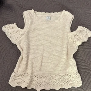 Beige stickad topp från Lindex - Säljer en söt beige stickad topp från Lindex med vackra spetsdetaljer och korta ärmar. Perfekt för en stilren och bekväm look. Toppen har en rund halsringning och är tillverkad i ett mjukt material.