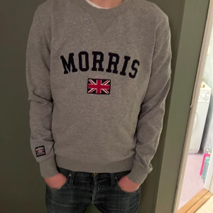Grå sweatshirt från Morris - Säljer en stilren grå sweatshirt från Morris med ett broderat brittiskt flaggmärke och text på framsidan. Tröjan har långa ärmar och ribbade muddar vid ärmslut och nederkant. Perfekt för en avslappnad stil.