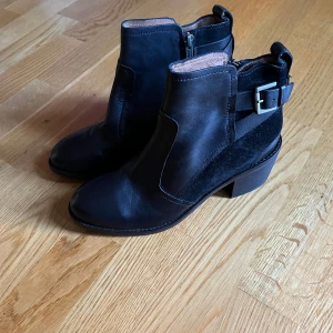 Svarta boots med spänne och dragkedja - Säljer ett par snygga svarta boots i skinn och mocka. De har en praktisk dragkedja på sidan och ett coolt spänne baktill. Perfekta för höst och vinter, och de är i storlek dam 38. Klacken ger en liten höjd utan att det blir obekvämt.