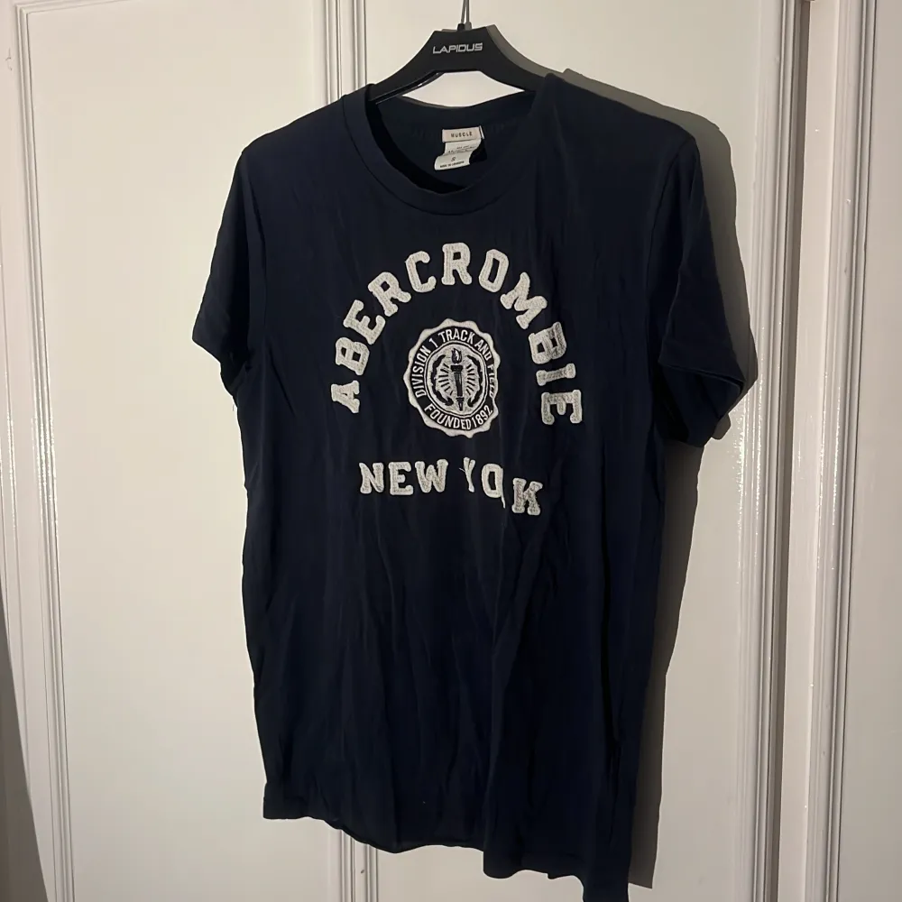 Säljer en snygg mörkblå t-shirt från Abercrombie & Fitch med texten 'Abercrombie New York' i vitt på framsidan. T-shirten är i storlek S och har en normal passform. Perfekt för en avslappnad stil!. T-shirts.