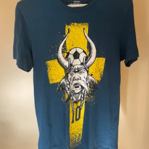 Blå t-shirt med vikingatema och nummer 10 - Rolig t-shirt med Sverige tema. Har ett tryck av en viking där fram å Sweden på baksidan. Kommer inte längre till användning. Skick 7/10