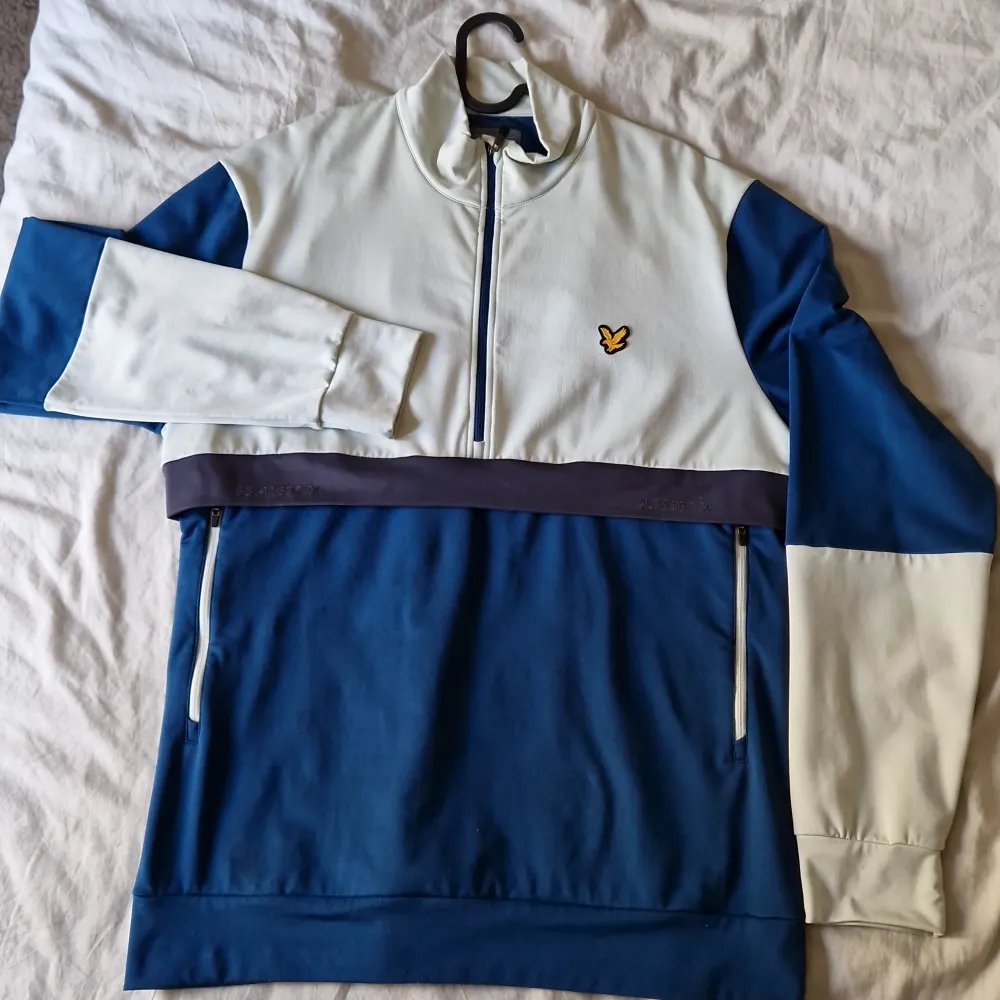 Säljer en snygg blå och vit tröja från Lyle & Scott. Den har en halv dragkedja framtill och ett coolt märke på bröstet. Tröjan är 88% polyester och 12% elastan, vilket gör den både bekväm och stretchig. Perfekt som träningströja men även vardag. . Tröjor & Koftor.