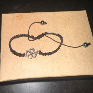Svart armband med fyrklöver - Säljer ett snyggt svart armband med en fyrklöver i silverfärg. Armbandet har en justerbar knytning med små pärlor i ändarna. Perfekt för att ge lite tur till din outfit!
