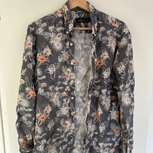 Blommig skjorta från Morris - Säljer en snygg blommig skjorta från Morris. Den har en mörk bas med vita och orange blommor som ger en cool och avslappnad look. Skjortan är långärmad och perfekt för både vardag och fest. Den är i bra skick och redo för en ny ägare!